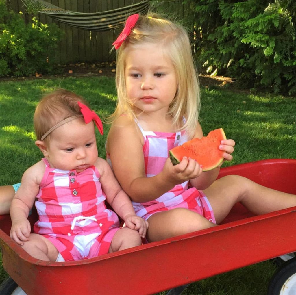 Infant and toddler sisters sitting in a vintage red radio flyer wagon wearing red and white checkered Carter's rompers eating watermelon in the summer. 