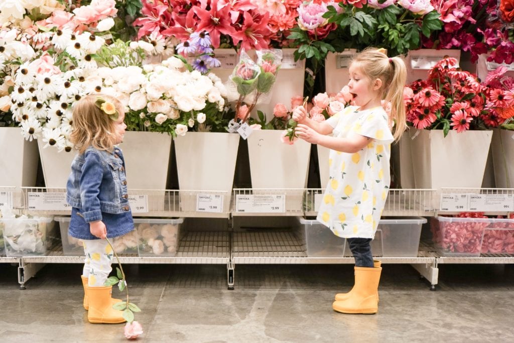 Toddler sisters wearing lemon spring clothing from Carters in matching yellow hunter boots at the floral section of Ikea. 