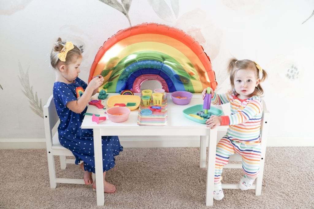 Toddler sisters playing with playdoh at a little white table with rainbow details in their Carter's rainbow pajamas. 