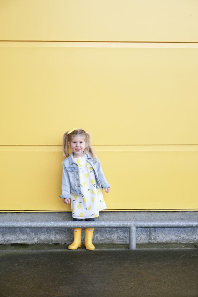 Toddler sisters wearing lemon spring clothing from Carters in matching yellow hunter boots in front of the yellow wall at Ikea. 