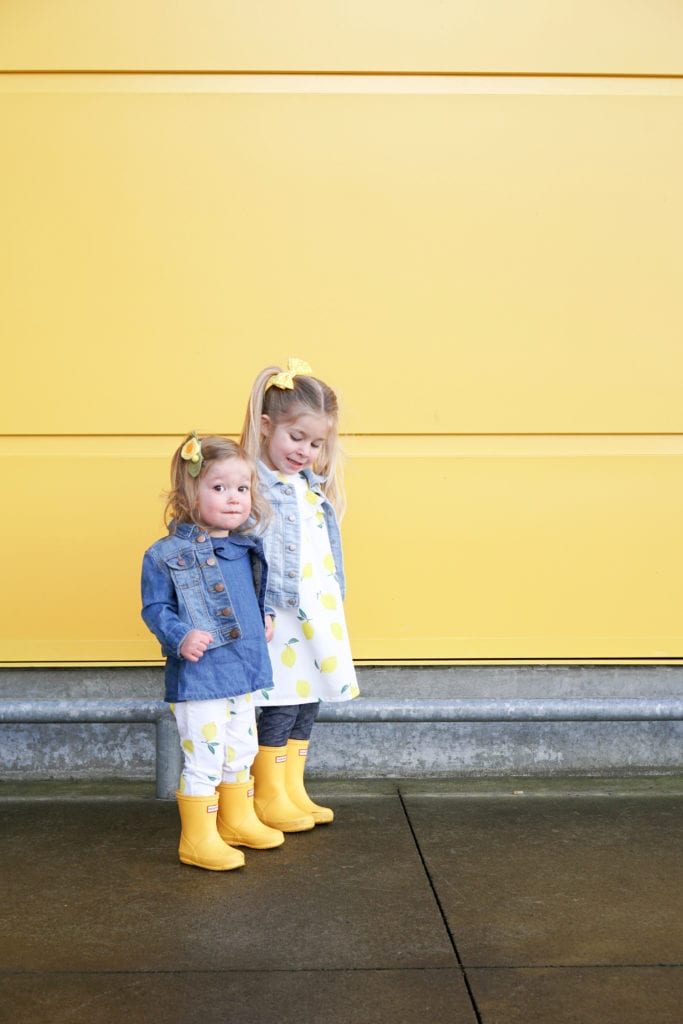Toddler sisters wearing lemon spring clothing from Carters in matching yellow hunter boots in front of the yellow wall at Ikea. 