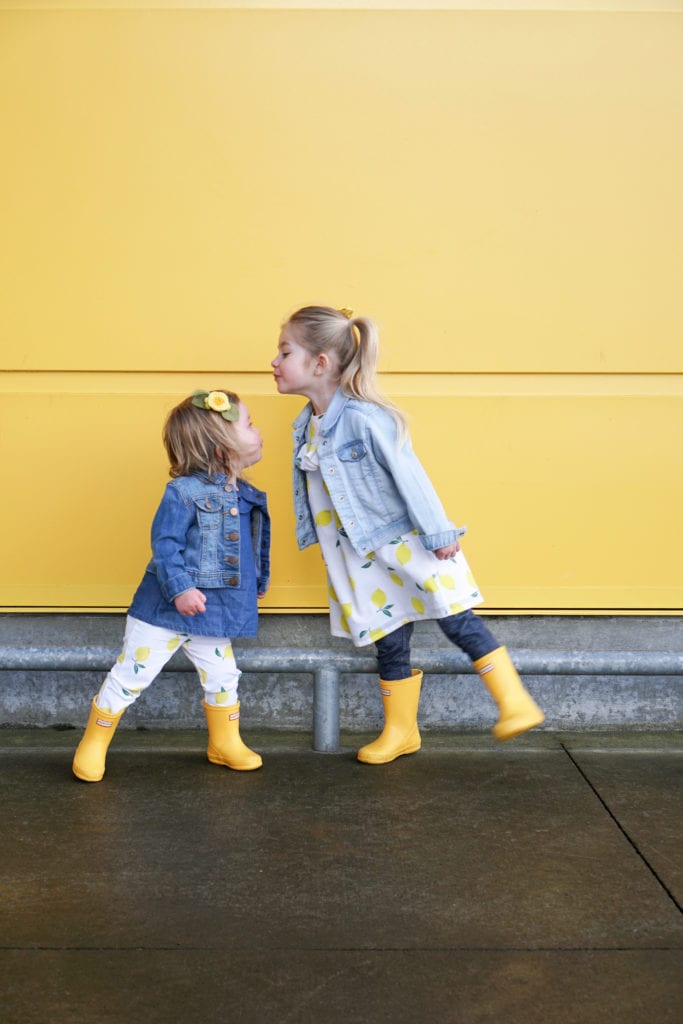 Toddler sisters wearing lemon spring clothing from Carters in matching yellow hunter boots in front of the yellow wall at Ikea. 