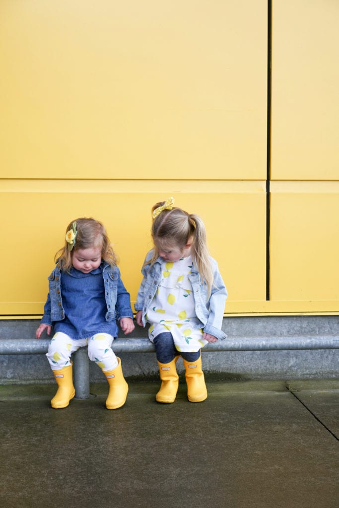 Toddler sisters wearing lemon spring clothing from Carters in matching yellow hunter boots in front of the yellow wall at Ikea. 