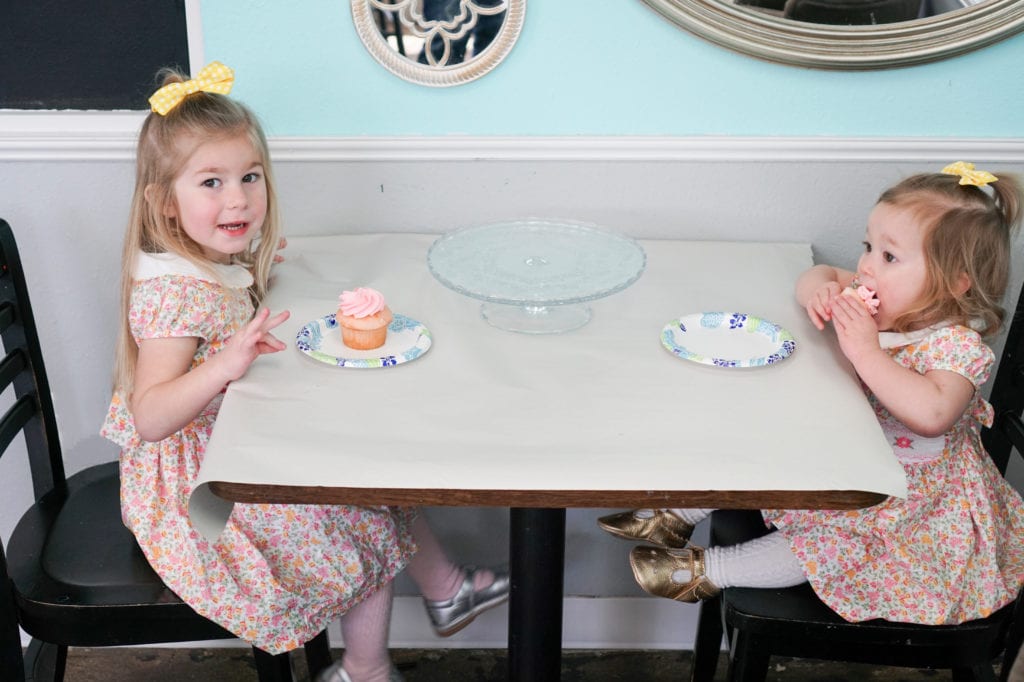 Toddler sisters in spring dresses eating pink cupcakes at a table. 